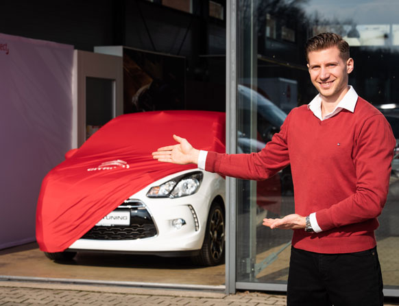 Fahrzeugübergabe – Mitarbeiter präsentiert weißes Auto unter rotem Tuch im Showroom