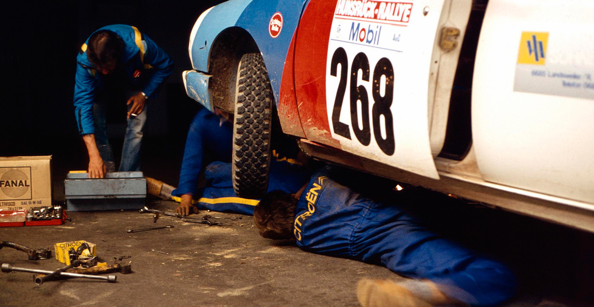 Citroën GSA Rallyefahrzeug mit der Startnummer 268 wird von Mechanikern in blauen Overalls gewartet. Einer arbeitet unter dem Fahrzeug, ein anderer steht daneben mit Werkzeugkasten und Ersatzteilen.