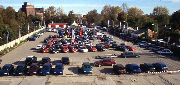 Großer Parkplatz mit Fahrzeugausstellung und Firmengebäude bei Sonnenschein