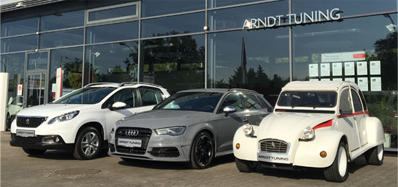 Firmengebäude Arndt Tuning Berlin – drei Fahrzeuge (SUV, Sportwagen, Oldtimer) vor Glasfassade mit Logo bei Sonnenschein