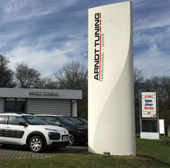 Gebäudeschild mit Logo und Fahrzeugen vor Firmengebäude bei Sonnenschein